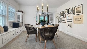 A dining area with a round table, four wicker chairs, a chandelier, and a wall with framed pictures and a sign.