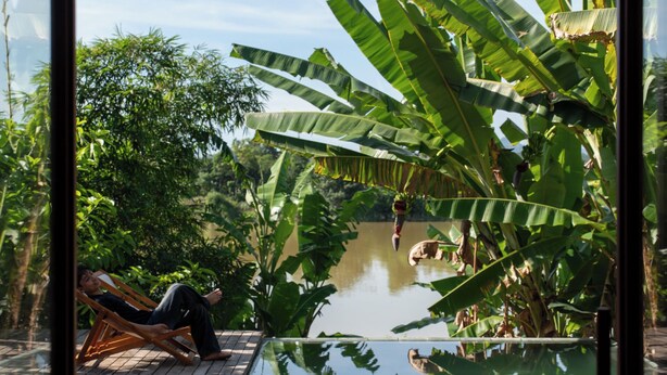 A glass-enclosed pool with a wooden deck, surrounded by lush greenery and banana plants. - Scarlet Pavilion Homestay (Hue)