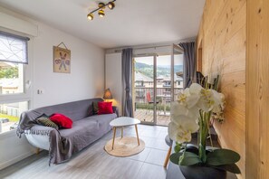 A modern living room with a grey sofa, a small white table, a lamp, and a view of a balcony with houses.
