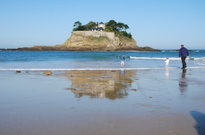 A beach with a rocky outcrop and a building on top, a person walking, and a child playing in the water.