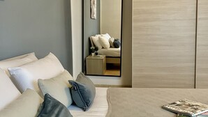 Een moderne slaapkamer met een bed, kussens, een nachtkastje met een lamp, en een spiegel die de kamer weerkaatst.
