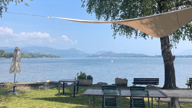 Een terras met tafels en stoelen, een overkapping en uitzicht op een meer met bergen op de achtergrond.