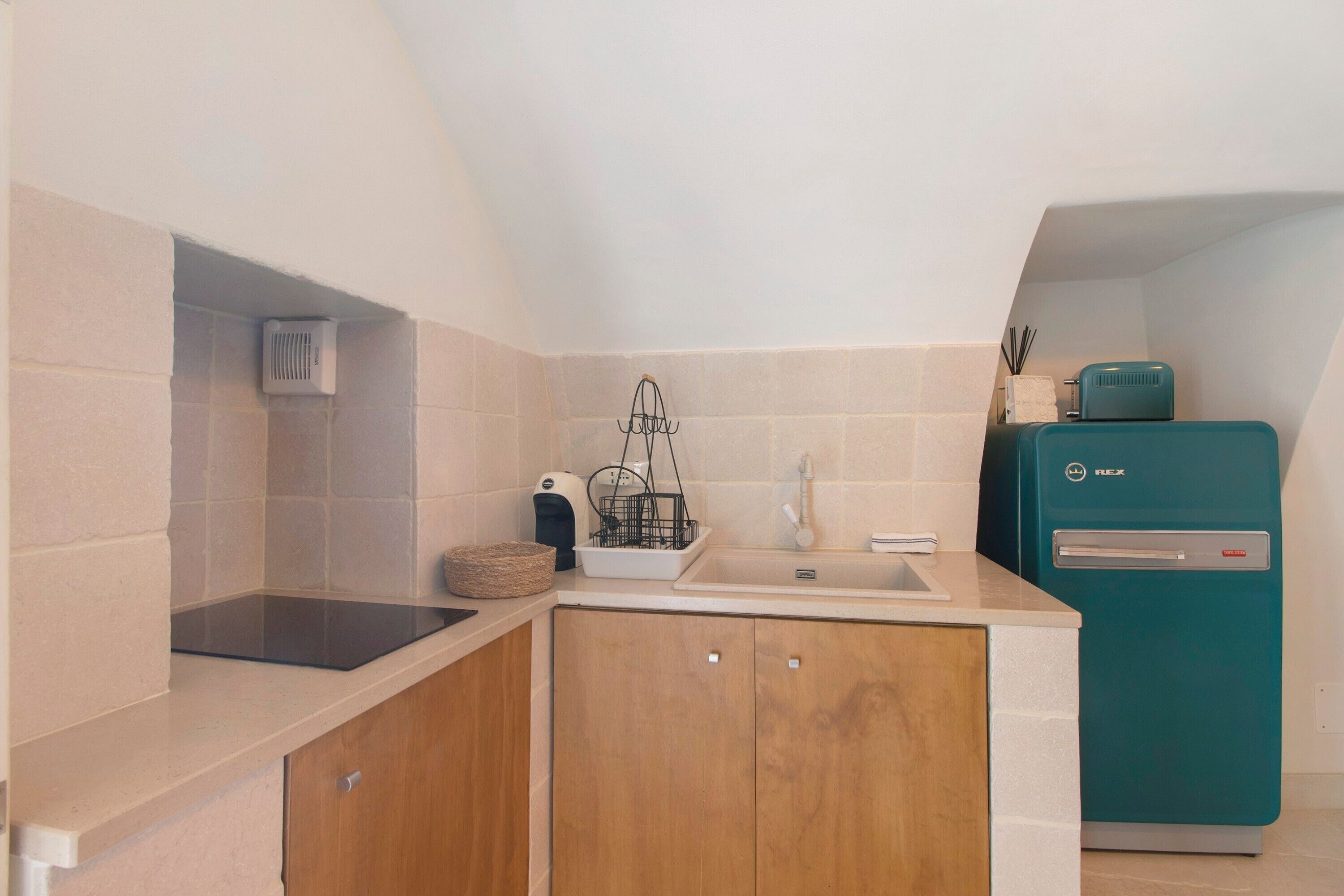 Une cuisine compacte dotée d’une cuisinière intégrée, d’un évier, de meubles en bois, d’un réfrigérateur de couleur turquoise et d’un petit îlot central avec un panier et une machine à café.
