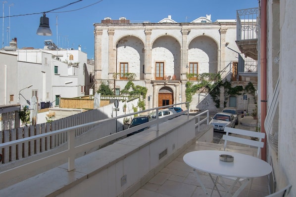 Una terraza con una mesa y sillas blancas, con vista a una calle con coches aparcados y un edificio histórico con arcos.