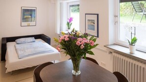 A bedroom with a bed, a bedside table, a window with a view of the outdoors, and a vase with flowers.