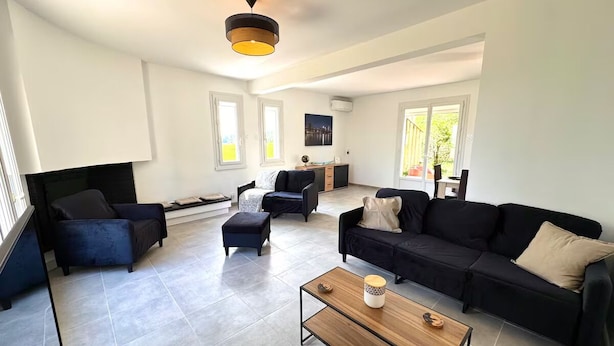 Uma sala de estar ampla com um sofá preto, uma poltrona azul e uma mesa de centro de madeira. Há duas janelas que proporcionam iluminação natural, e um ventilador de teto é visível.