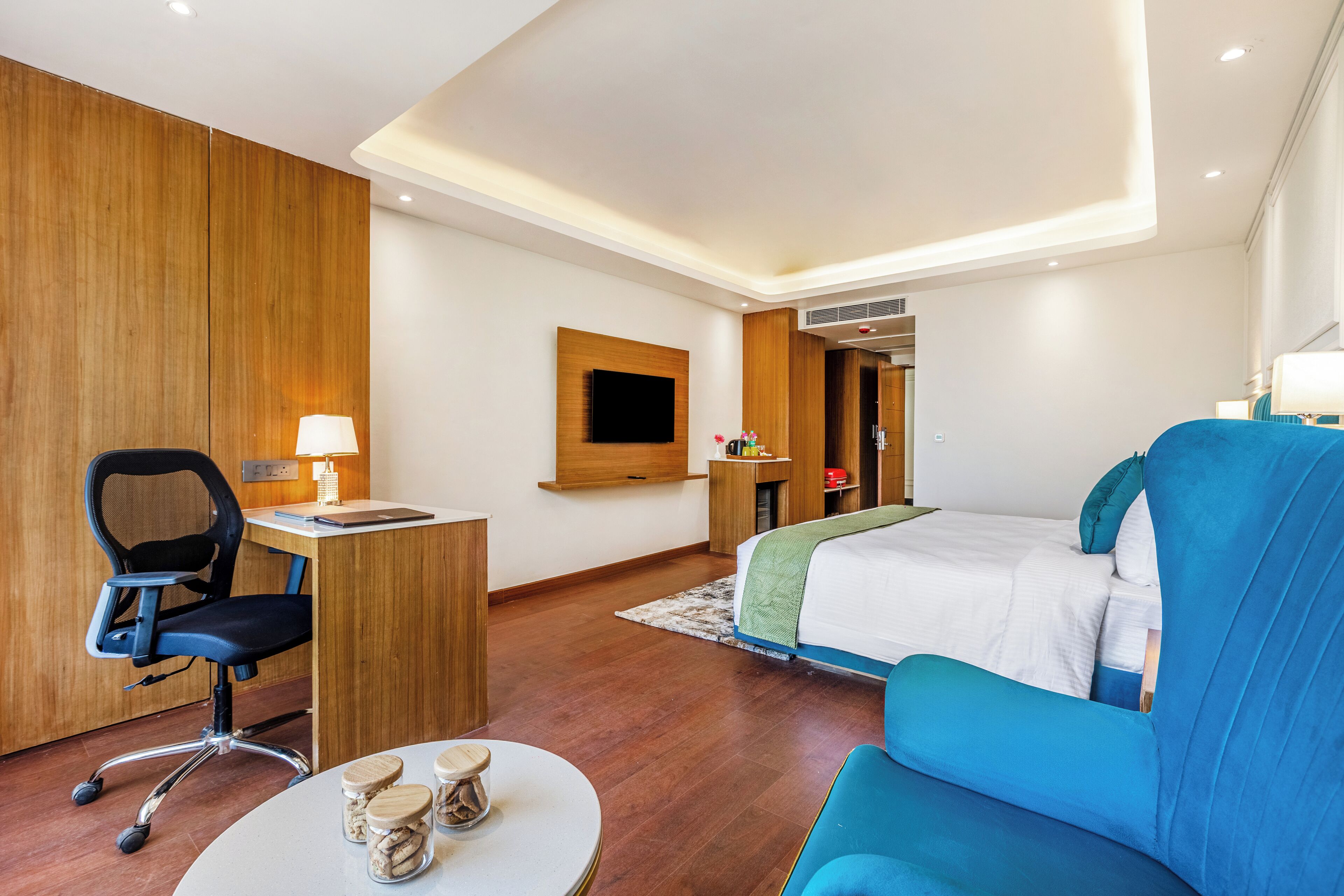 Una habitación de hotel con cama, escritorio con silla, televisor montado en la pared y un sillón azul.