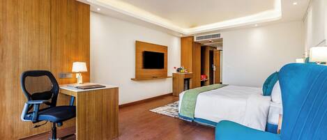Una habitación de hotel con cama, escritorio con silla, televisor montado en la pared y un sillón azul.