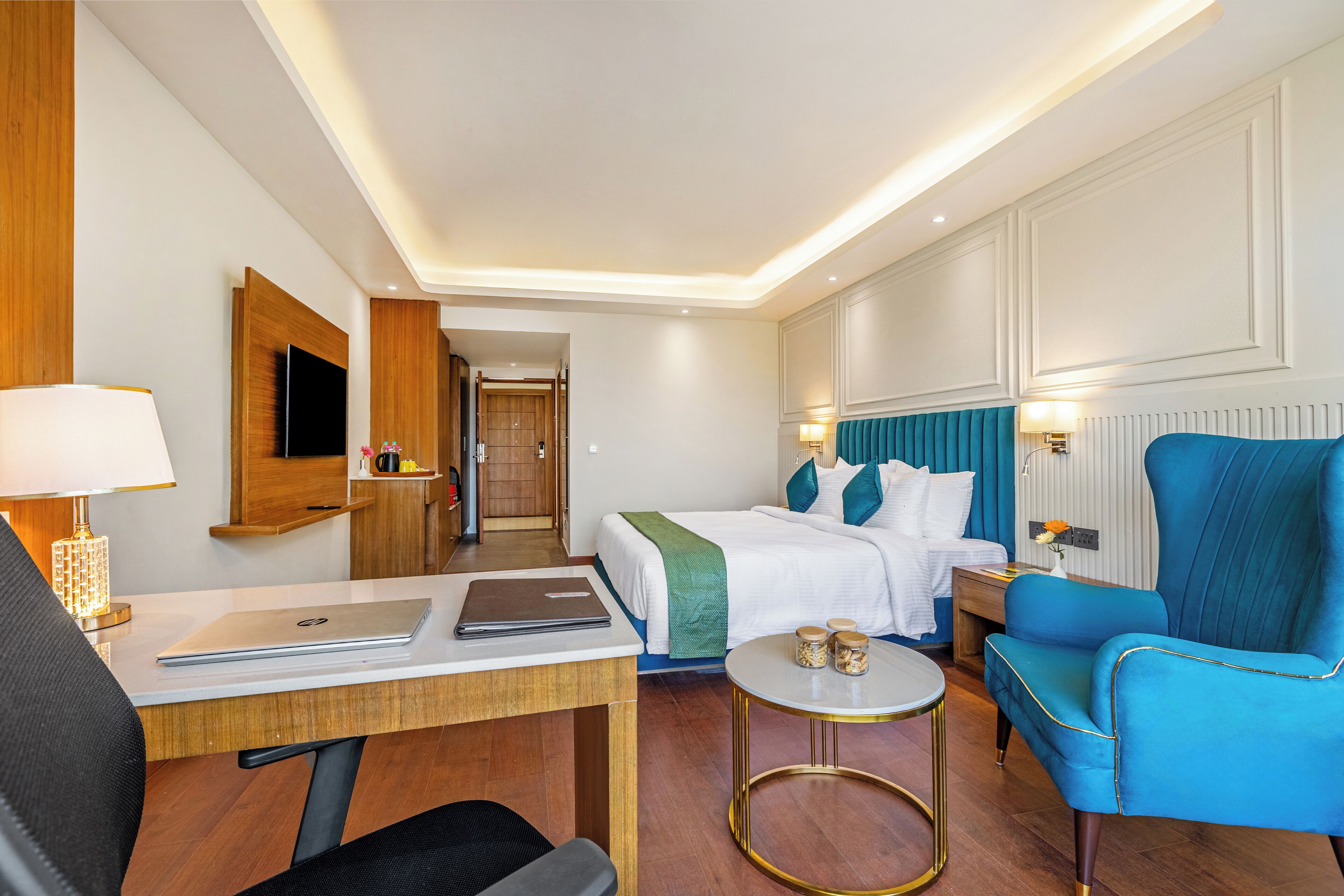 A hotel room with a bed, a desk with a laptop, a blue armchair, and a small round table with jars.