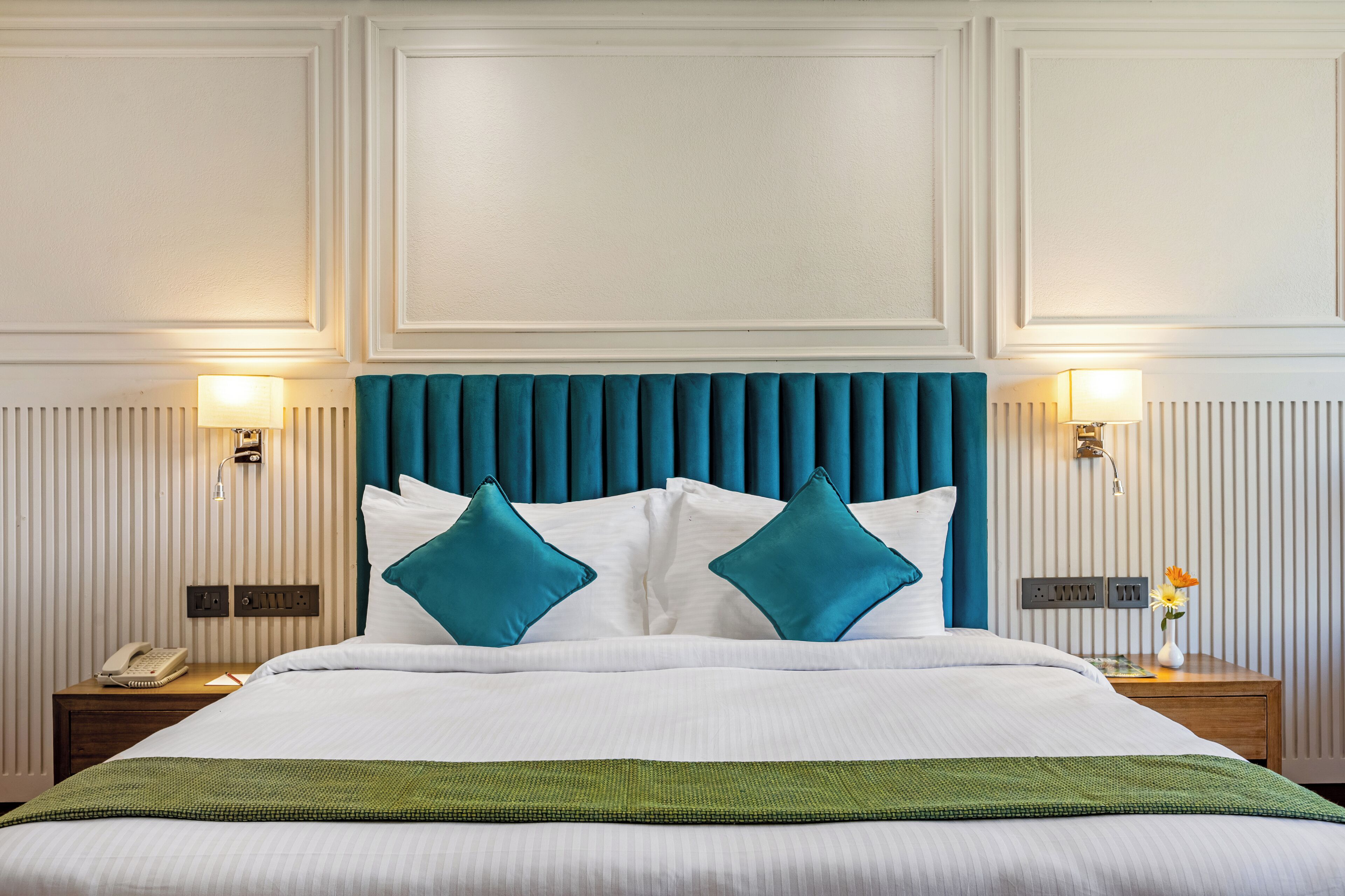 A neatly made bed with a teal headboard, white bedding, and two teal pillows. There are bedside lamps, a telephone, and a small table with a vase of flowers.
