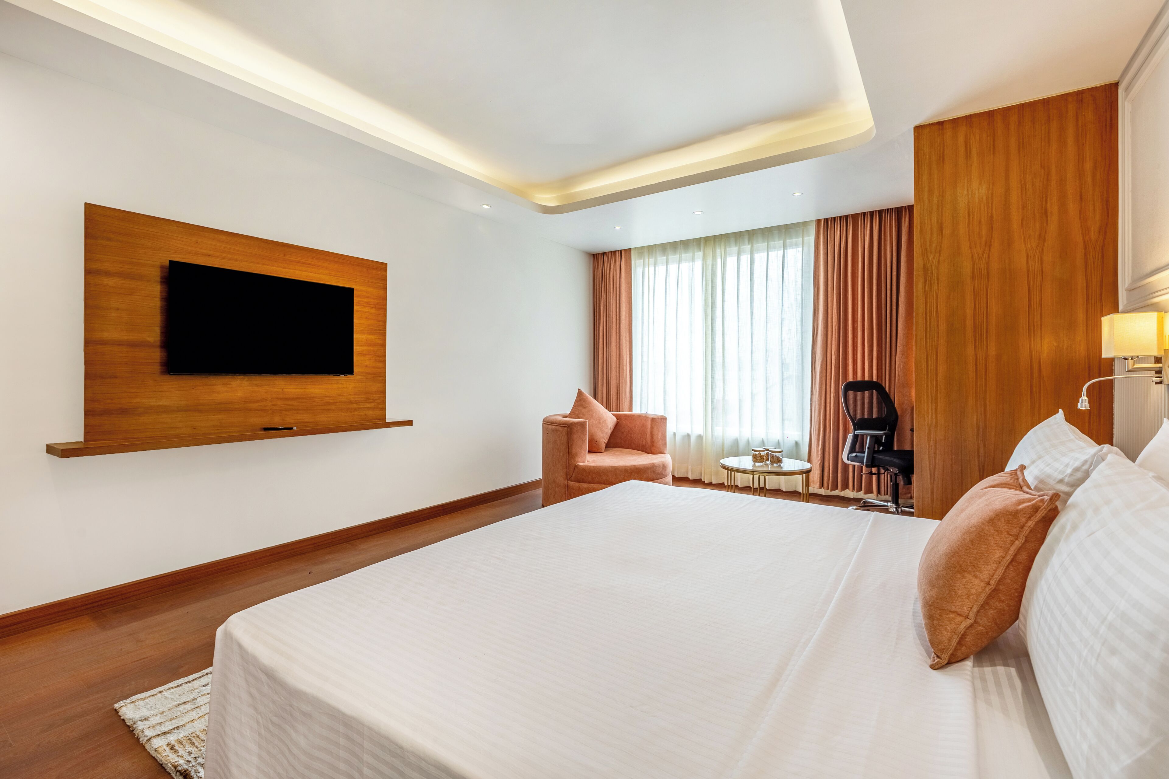 A modern hotel room with a large bed, a flat-screen TV mounted on the wall, a small table, a chair, and a window with curtains.