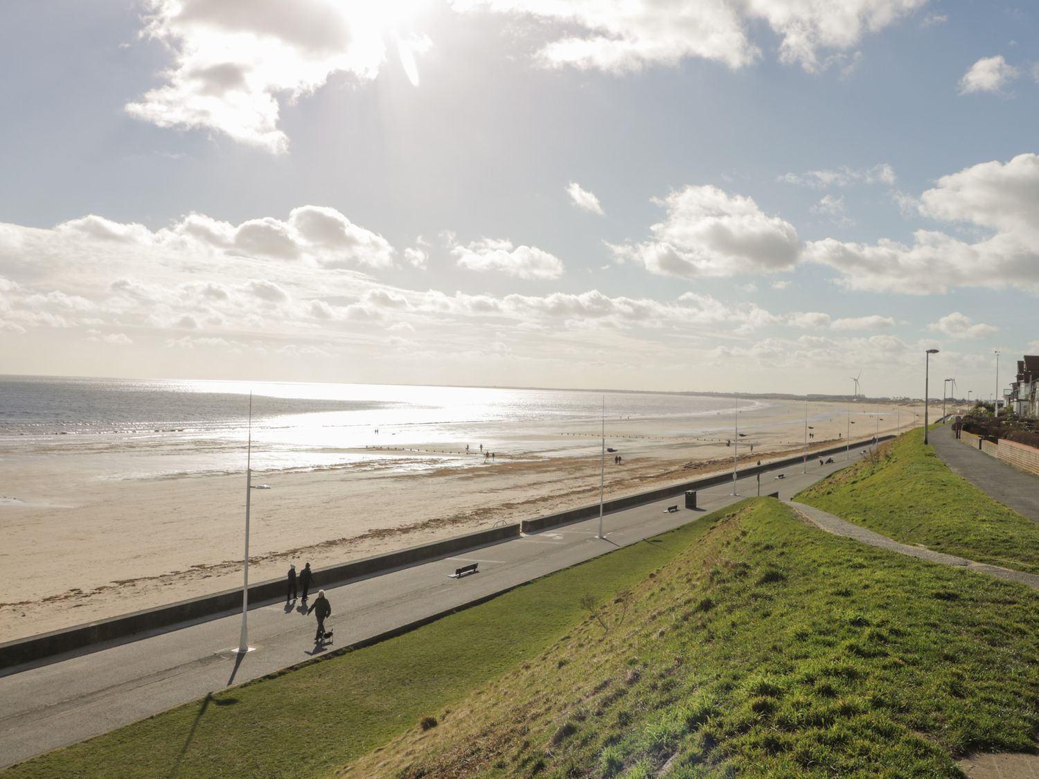 Eine Küstenstraße mit einer grasbewachsenen Böschung, einem Fußweg und einem Strand im Hintergrund.