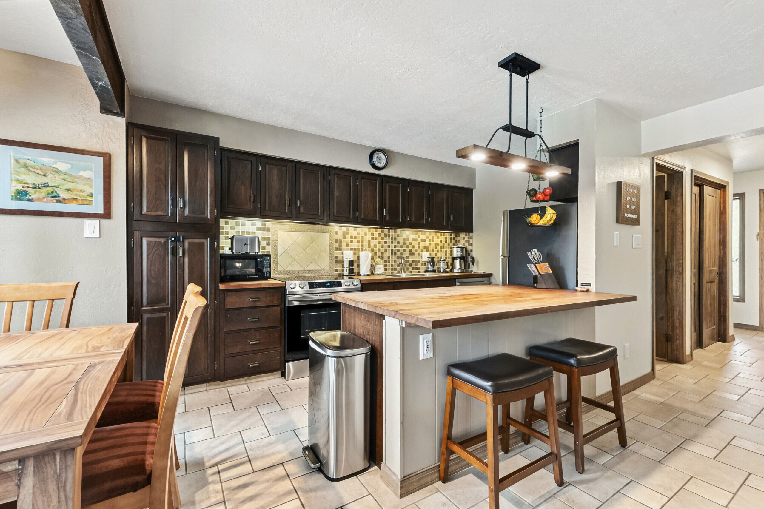 A modern kitchen with dark wooden cabinets, a large island with stools, a stainless steel trash can, and a clock on the wall.