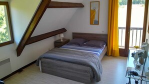 Un dormitorio con techo inclinado, una cama con ropa de cama a rayas, una mesita de noche con una lámpara, un cuadro en la pared y un balcón con cortinas amarillas.