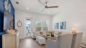 Ein modernes Wohnzimmer mit einer Sofagarnitur, Hocker und einem Couchtisch. Es gibt einen Deckenventilator, einen wandmontierten Spiegel und ein Fenster mit Jalousien.