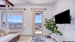 A modern living room with a sofa, a flat-screen TV, a large window with curtains, a potted plant, and a view of a cityscape. - Apt Smode With Panoramic Balcony (Koper)