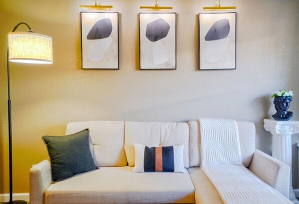 A modern living room with a sofa, three framed abstract paintings, a floor lamp, and a vase with flowers.