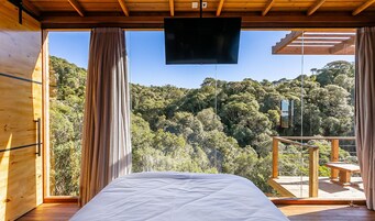 A bedroom with a bed, wooden walls, a TV, and a balcony with a view of a forested area.