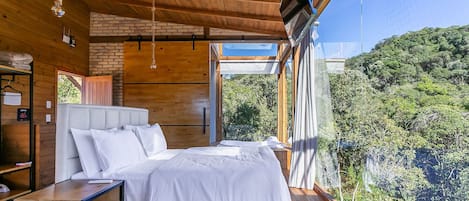 A bedroom with a large bed, wooden ceiling, and a view of a forested hillside.