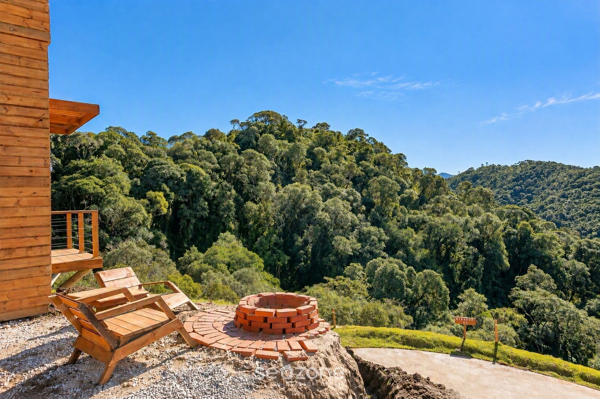 A wooden deck with lounge chairs and a brick fire pit, overlooking a dense forest and hills.