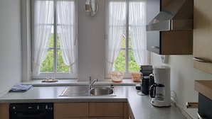 Una cocina con un lavamanos de acero inoxidable, una cafetera y dos ventanas con cortinas translúcidas.