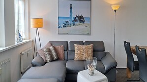 Un salón moderno con un sofá gris, una mesa de centro de cristal, un cuadro de un faro y una lámpara de pie.