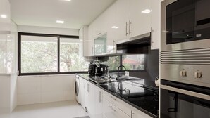 A modern kitchen with white cabinets, a black countertop, a built-in oven, and a window with a view of trees. - The Light Stay at Cascais (Cascais)