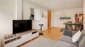 A modern living room with a flat-screen TV, a grey sofa, a wooden entertainment unit, and a dining area with a table and chairs. - The Light Stay at Cascais (Cascais)