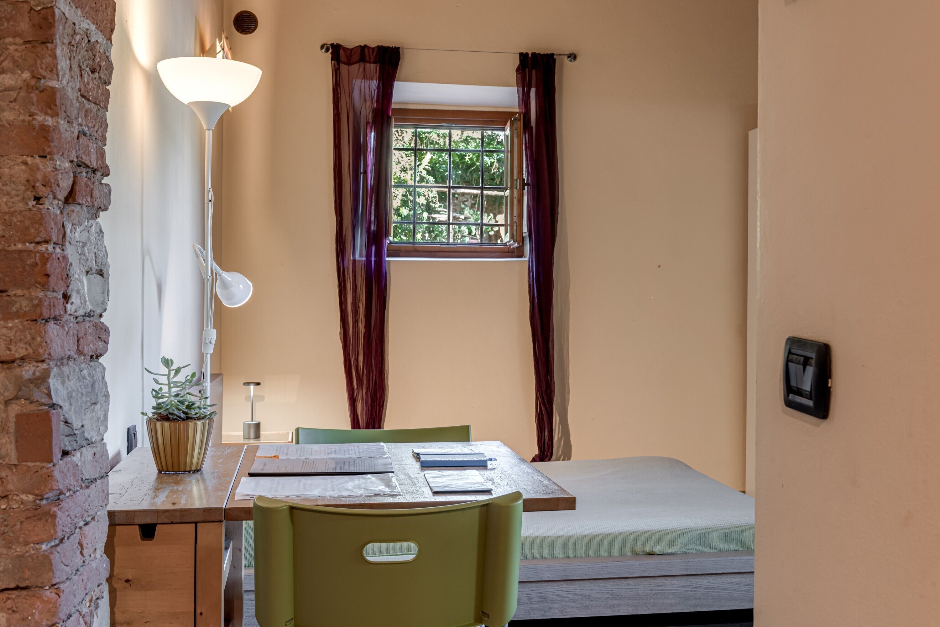 A small room with a bed, a desk with a lamp, a chair, and a window with curtains.