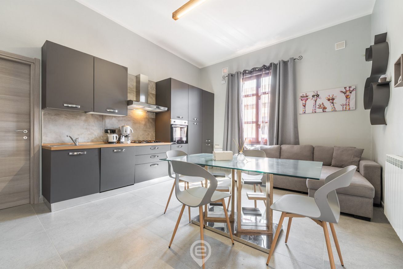 A modern kitchen with dark cabinets, a glass dining table, and a grey sofa in the living area.