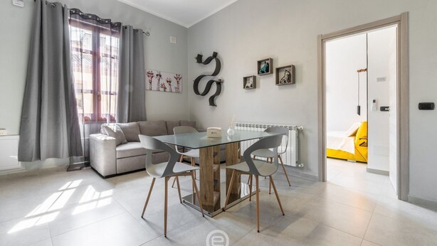 Un salon moderne avec une table à manger en verre, un canapé gris et un lit jaune dans la pièce adjacente.