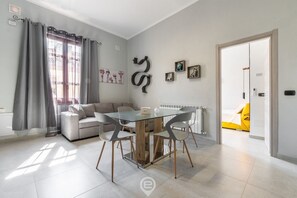 A modern living room with a glass dining table, grey sofa, and a yellow bed in the adjacent room. - Cagliari Shopping District Flat (Cagliari)