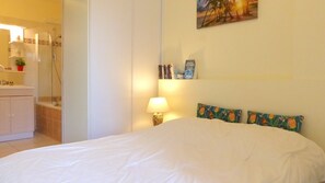 A bedroom with a bed, a nightstand with a lamp, a bookshelf, and a bathroom visible in the background. - Cosy flat close to the centre of Auray (Auray)