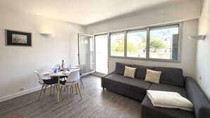 A modern living room with a grey sofa, a dining table with chairs, and a large window offering a view of buildings and trees. - Jetée des Marins by Interhome (Arcachon)