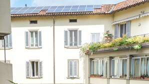 Un balcon avec une table en bois et une chaise rouge, donnant sur un immeuble doté de nombreuses fenêtres et d’un toit recouvert de tuiles.