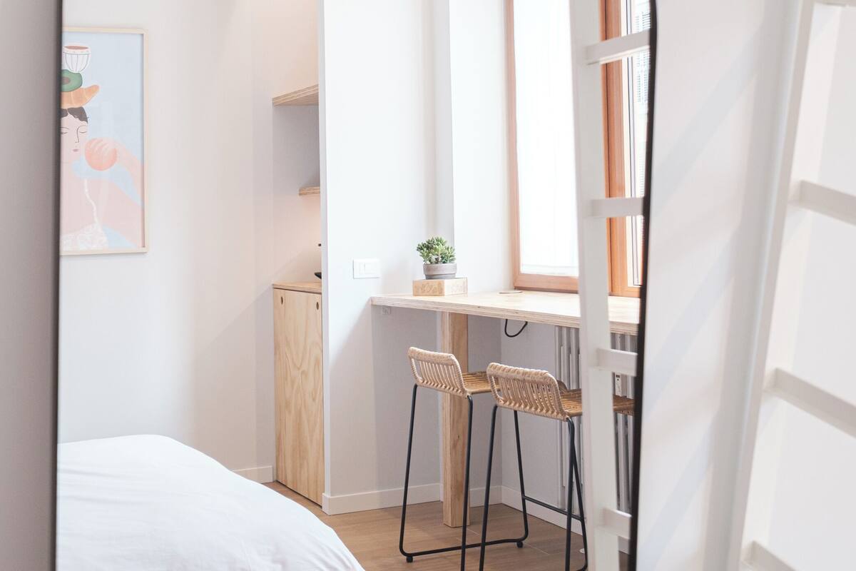 A narrow room with a bed, a desk with two chairs, a window, and a door.