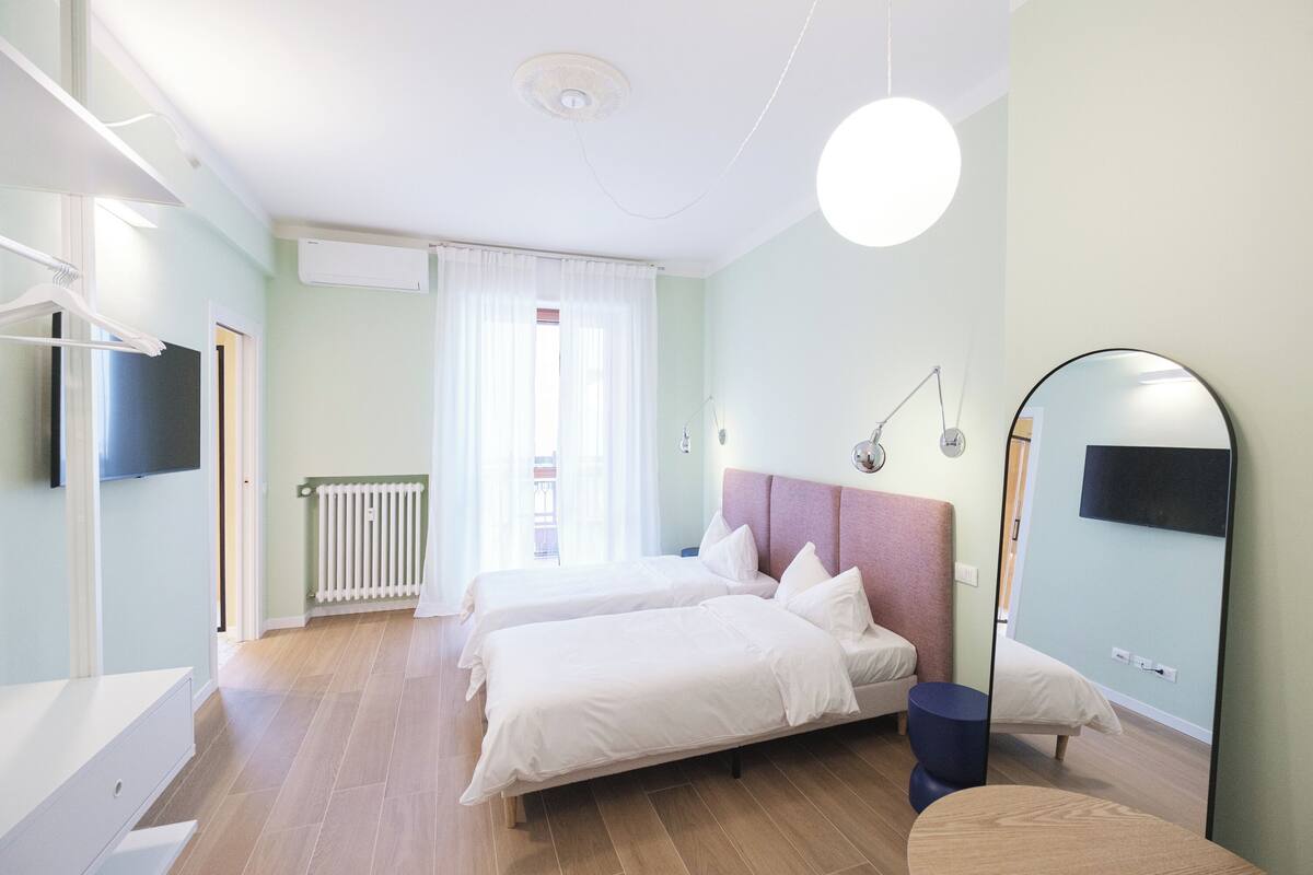 A modern bedroom with two beds, a large mirror, a bedside table, and a hanging light fixture.