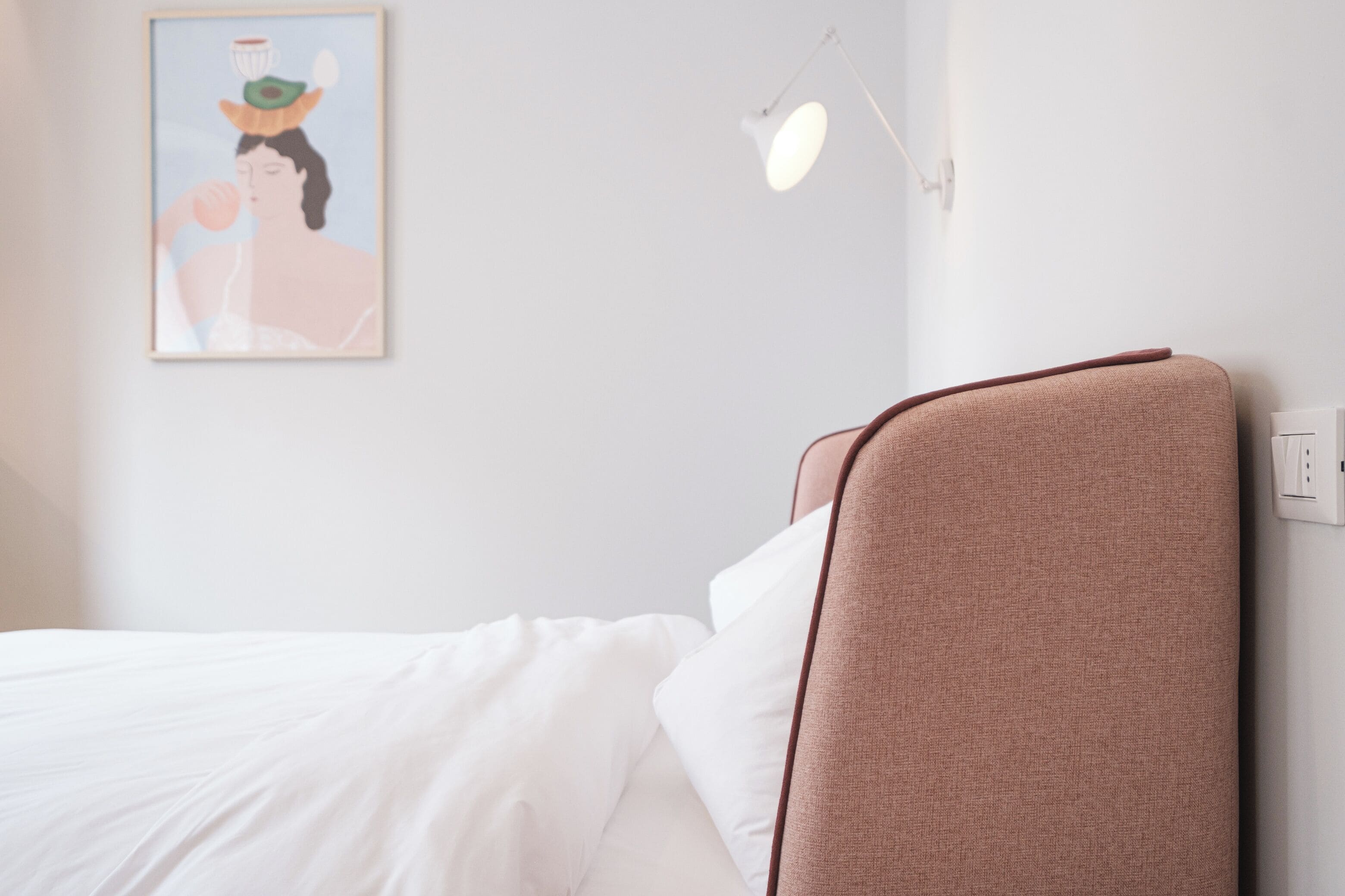 A neatly made bed with white linens, a headboard with a brown fabric, and a wall-mounted light fixture.