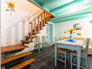 A staircase with wooden steps and a wooden railing, leading to a room with a table and chairs, a sofa, and a wall with a sunflower decoration.