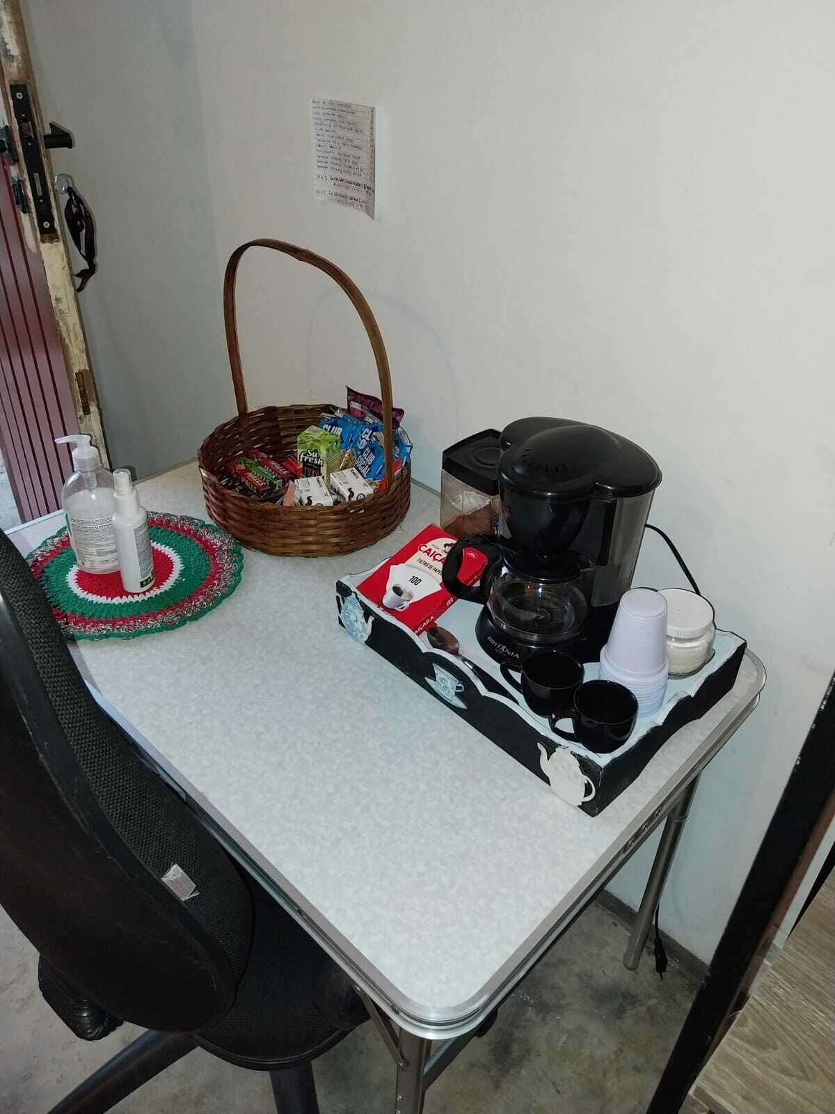 A small kitchenette with a coffee maker, cups, a basket of snacks, and a bottle of hand sanitizer on a white countertop.
