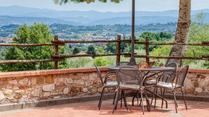 A patio with stone walls, metal chairs, and a table overlooking a scenic landscape with mountains and greenery.