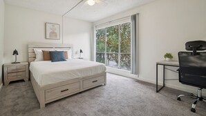 Une chambre à coucher comprenant un lit, un bureau, une chaise, un ventilateur de plafond et une fenêtre donnant sur des arbres.