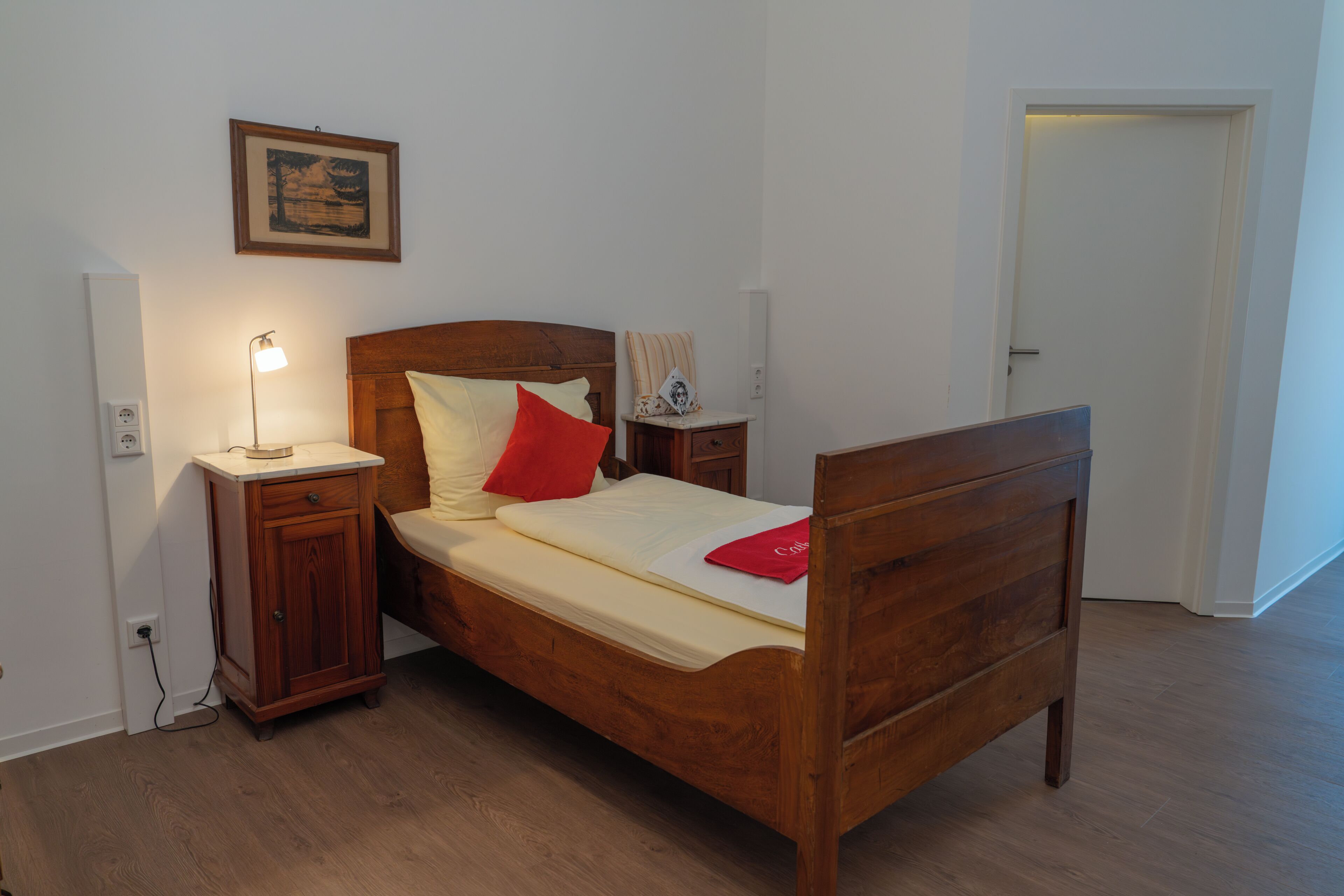 A wooden bed with a headboard, a bedside table with a lamp, a nightstand, and a framed picture on the wall.