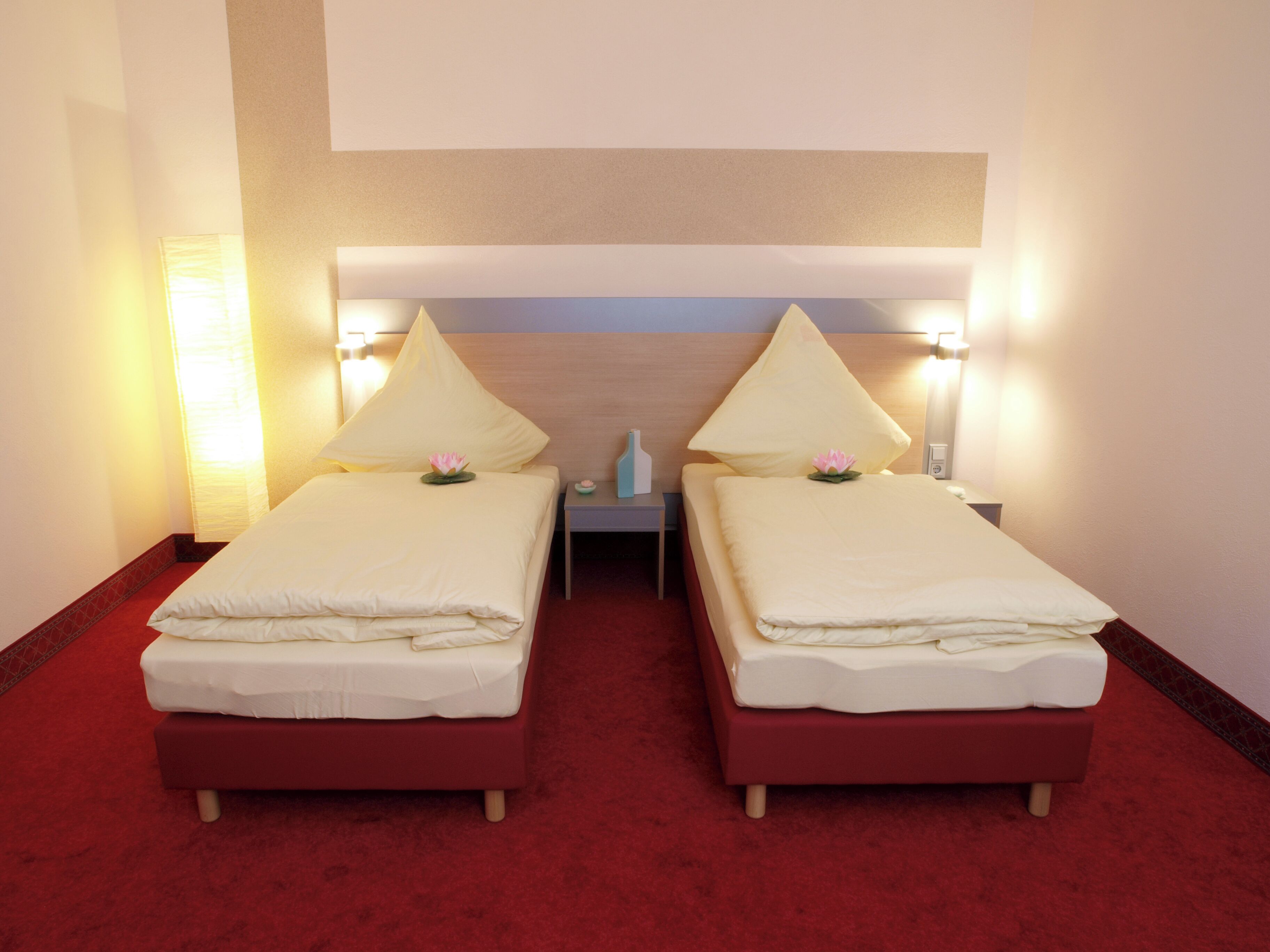 A hotel room with two beds, each with a pillow and a flower. There is a nightstand between the beds with a lamp and a small vase. The room has a red carpet and a wooden headboard.