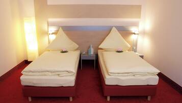 A hotel room with two beds, each with a pillow and a flower. There is a nightstand between the beds with a lamp and a small vase. The room has a red carpet and a wooden headboard.