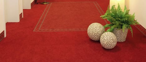 Pasillo de hotel con alfombra roja, varias puertas y lámparas fijadas a la pared.
