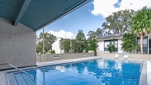 A rectangular swimming pool with clear blue water, surrounded by a concrete deck. There are lounge chairs and a palm tree visible in the background.