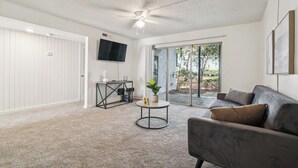 A spacious living room with a grey sofa, a round coffee table, a ceiling fan, and a sliding glass door leading to a patio.
