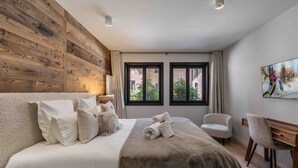 A hotel room with a wooden headboard, a large bed with multiple pillows, a desk with a chair, and a window with a view of buildings and trees. - Modern duplex near slopes with parking (Courchevel 1550)