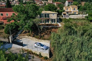 Uma casa de dois andares com um carro estacionado na entrada, cercada por vegetação e outras casas.